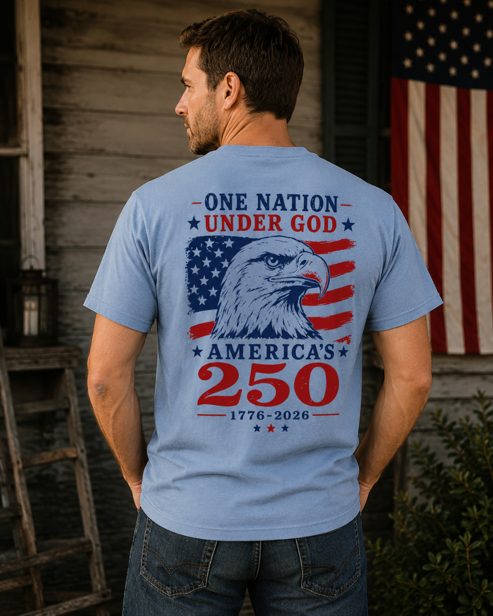 Man wearing a blue t-shirt with an American flag and eagle design, standing in front of a wooden building and American flag.